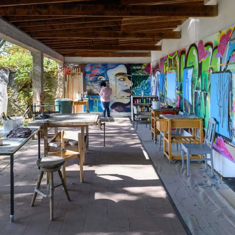 Überdachter Außenbereich mit Tischen und Stühlen, an der Wand hängen bunte Gemälde, im Hintergrund sind Bäume sichtbar.